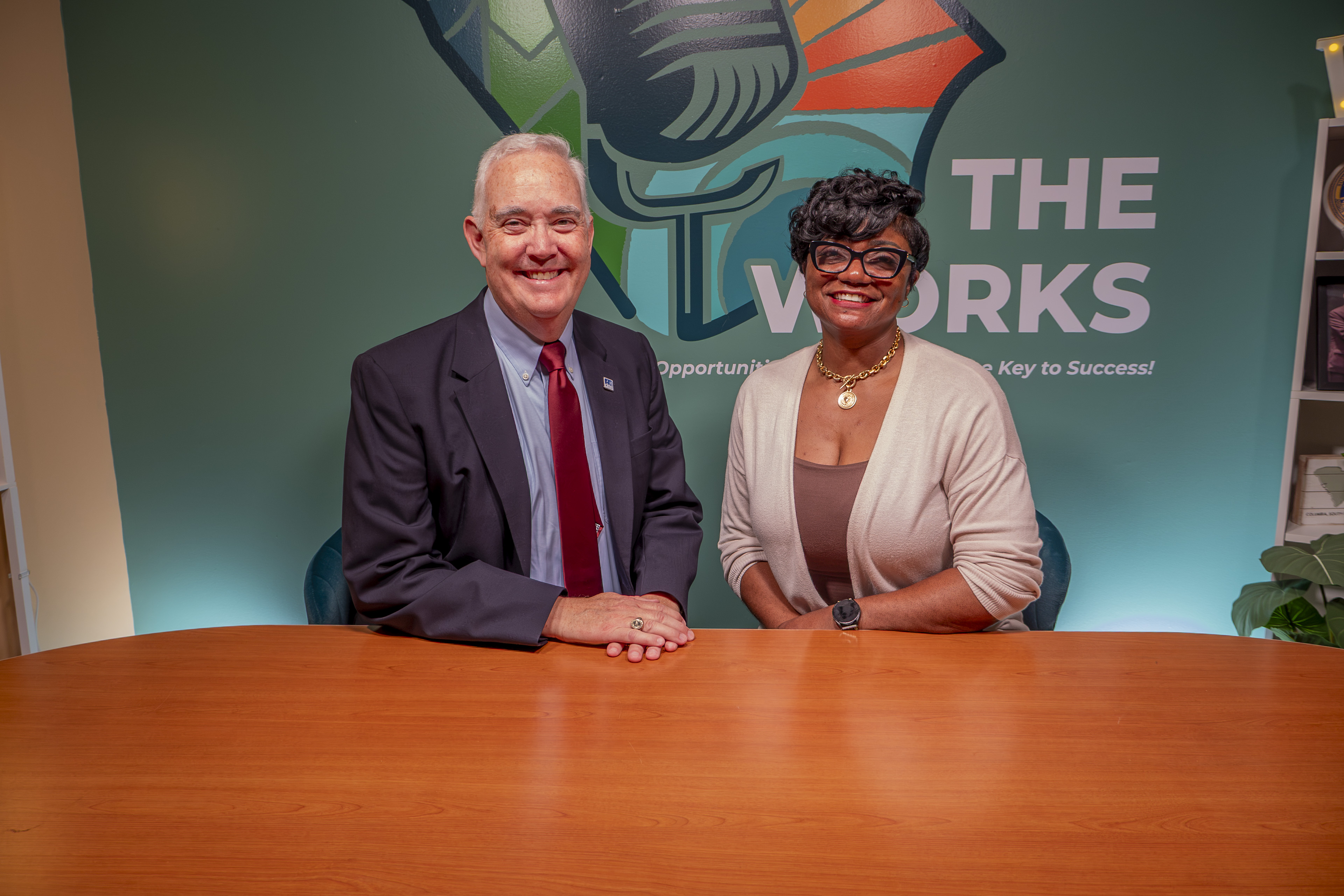 A man and a woman sit at the table, both are smiling, there is the Works logo behind them. 