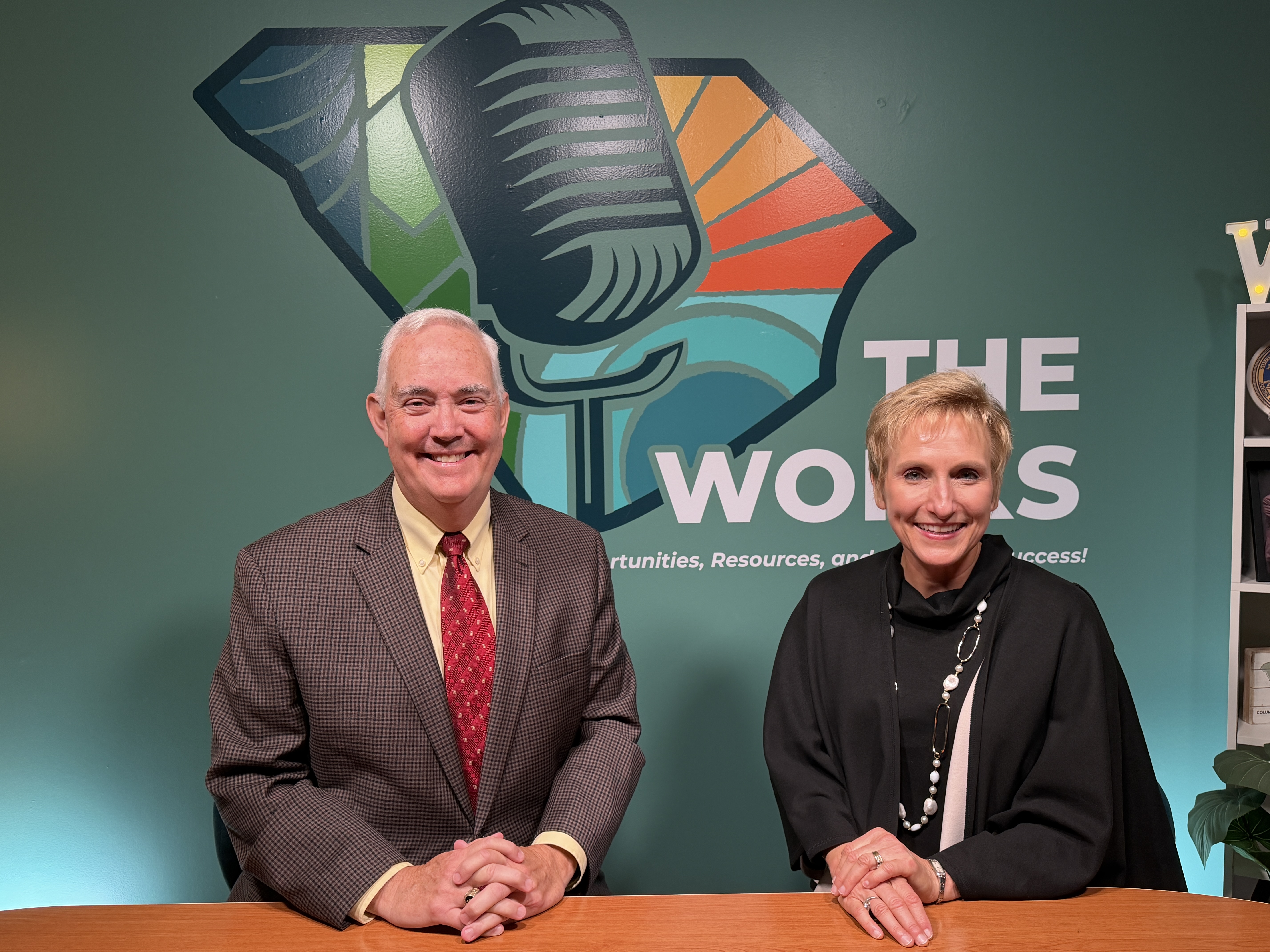 A man in a brown suit with a red tie is smiling next to a woman with a black jacket on. She is also smiling.