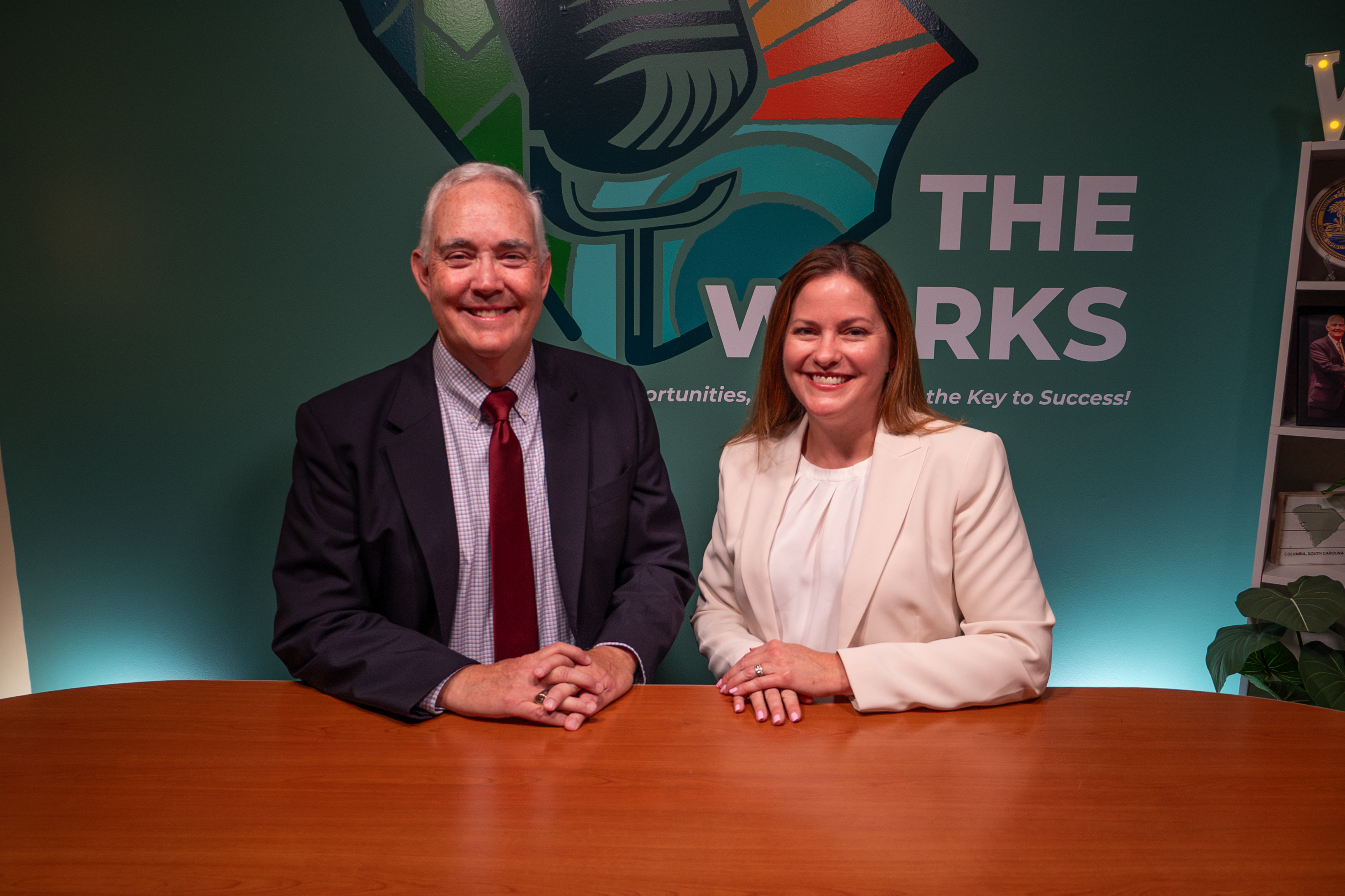 A man and a woman are seated next to each other at a table. They are smiling. There is a logo for the Works podcast in the background.
